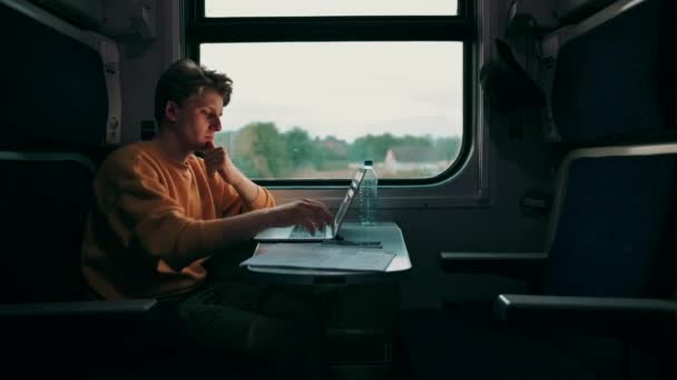 Bonito jovem monta à noite em um compartimento de trem em uma mesa com um laptop e tira fotos da janela em uma câmera de smartphones. Guy usa Internet em gadgets enquanto viaja de trem. - Filmagem, Vídeo