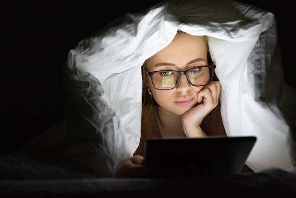 Vrouw liggend in bed onder de deken met behulp van tablet 's avonds laat, kan niet slapen, nieuws zoeken over Covid-19 of scrollen sociale netwerken. Slapeloosheid, nomofobie, slaapstoornis.   - Foto, afbeelding
