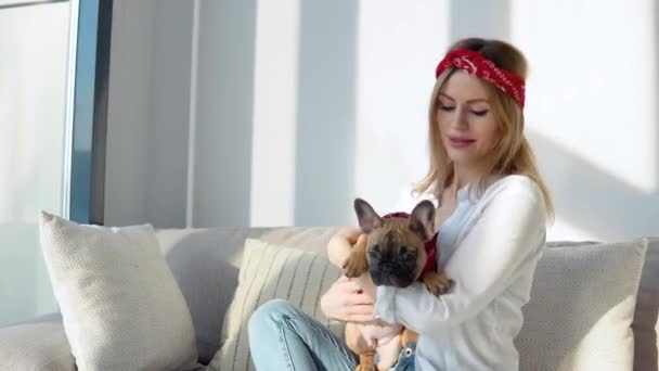 Mujer joven en una camisa blanca y jeans se sienta en el sofá y juega con su perro - Metraje, vídeo