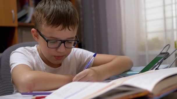Lächelnder Schuljunge mit Brille schreibt mit Stift ins Notizbuch, spricht mit dem Smartphone - Filmmaterial, Video