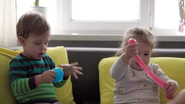 auténticas manos de bebé con limo azul y rojo. Niña y niño preescolar menor estiramiento Play-Doh a los lados. niño jugando plastilina. Juguete líquido de moda se pega a las manos y los dedos en un sofá gris amarillo - Imágenes, Vídeo