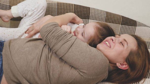 Little child plays with mommy and laughs while lying on the couch. Happy girl rejoices at mom. A daughter and a woman are playing in the childrens room. Mothers Day. Parents love for kid. Upbringing - Photo, Image