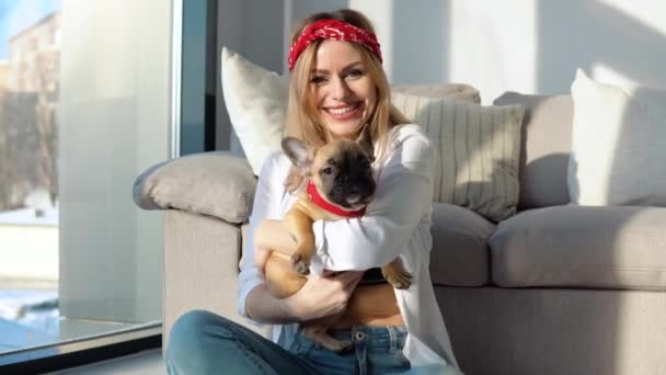 Young woman in a white shirt and jeans sits on the floor near the sofa with her small french bulldog - Footage, Video