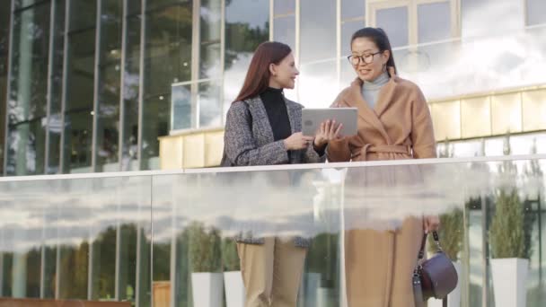 Низкоугольный средний снимок дружелюбных многоэтнических женщин-коллег с цифровым ноутбуком, стоящих снаружи футуристического офисного здания, говорящих о работе - Кадры, видео