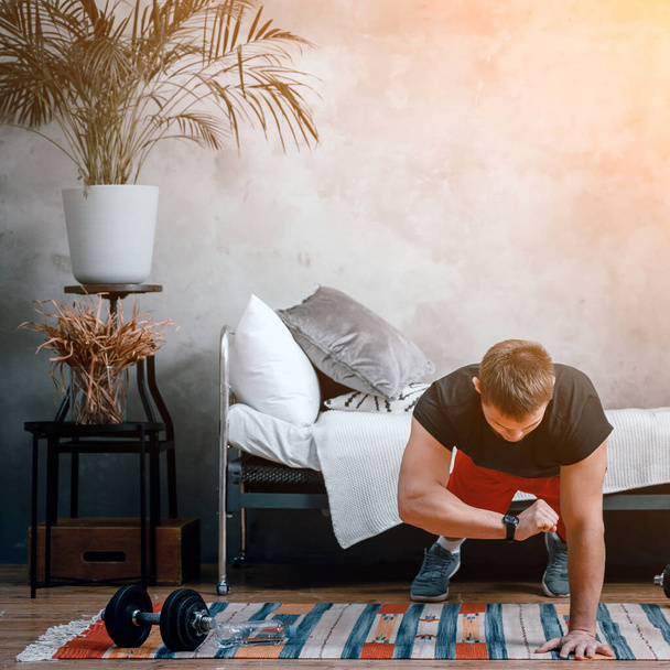 Jongeman gaat thuis sporten en traint online. De atleet maakt een plank, kijkt naar de tijd op een sporthorloge, een stopwatch in de slaapkamer, op de achtergrond een bed, een vaas, een tapijt. - Foto, afbeelding