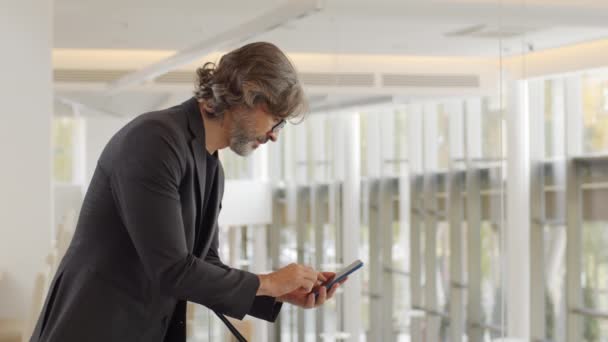 Slow-motion side-view střední záběr kavkazského dospělého podnikatele pomocí smartphonu nakloněného přes zábradlí ve druhém patře moderní budovy - Záběry, video
