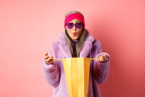 Compras y concepto de moda. Elegante mujer anciana asiática en gafas de sol y abrigo de piel sintética bolsa de papel abierta con regalos, mirando sorprendido a la cámara, fondo rosa - Foto, Imagen