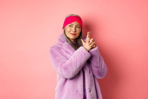 Mode- und Einkaufskonzept. Coole und stylische asiatische Seniorin im lila Kunstpelzmantel, mit Fingergewehr-Geste und einem frechen Lächeln nach links schauend, wie ein Geheimagent agierend, rosa Hintergrund - Foto, Bild