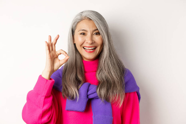 Detailní záběr krásné asijské dospělé ženy ukazující OK znamení a usmívající se, schválit a podobný produkt, stojí spokojeni s bílým pozadím - Fotografie, Obrázek