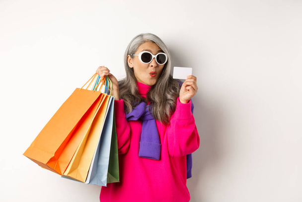 Modaya uygun Asyalı yaşlı kadın alışverişe gidiyor, elinde kağıt torbalar ve plastik kredi kartı var, şık güneş gözlüğü takıyor, beyaz arka planda duruyor. - Fotoğraf, Görsel