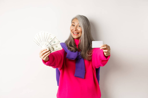 Ostokset käsite. Iloinen aasialainen vanhempi nainen, joka näyttää rahaa ja muovista luottokorttia, hymyilee kameralle, seisoo valkoisen taustan yläpuolella - Valokuva, kuva