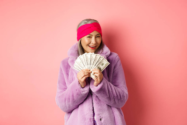 Shopping and fashion concept. Fashionable asian senior woman thinking about buying new clothes, showing money in dollars and smiling greedy, pink background - Photo, Image