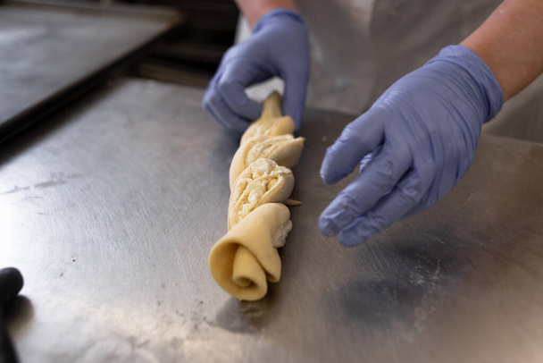 Τα χέρια του μάγειρα σε μπλε προστατευτικά γάντια ετοιμάζουν ένα ρολό γεμιστό με τυρί cottage και ζάχαρη. Χειροκίνητη παραγωγή προϊόντων αλεύρου. - Φωτογραφία, εικόνα