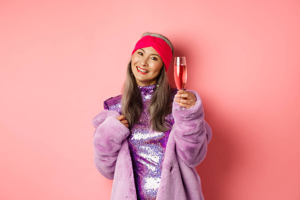 Hermosa asiática senior hembra en elegante abrigo de piel sintética púrpura y vestido brillante, levantando la copa de champán en el gesto de vítores, diciendo brindis en la fiesta, fondo rosa - Foto, Imagen