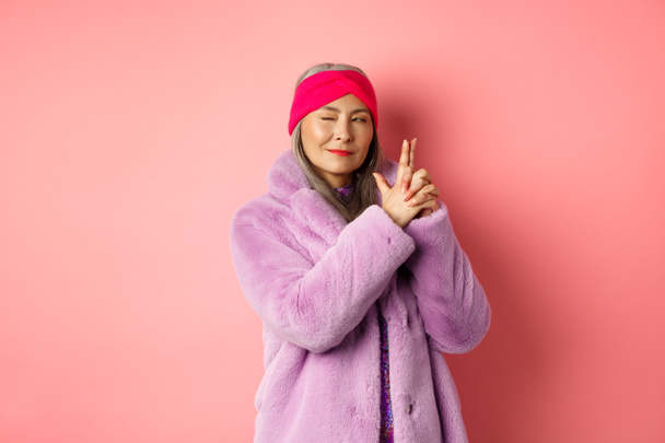 ファッションとショッピングのコンセプト。クールでスタイリッシュなアジアのシニア女性で紫偽の毛皮のコート,指銃ジェスチャーを作り、おしゃれな笑顔で左を見ます,秘密のエージェントのように機能します,ピンクの背景 - 写真・画像