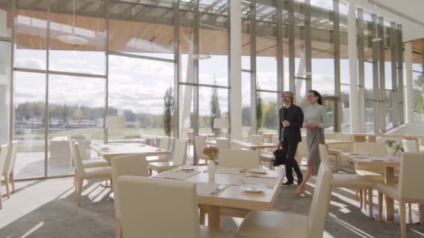 Panning śledzenie side-view slow-motion pełne ujęcie wieloetnicznej pary partnerów biznesowych spacerujących wzdłuż luksusowej restauracji znajduje się na pierwszym piętrze nowoczesnego budynku z oknami panoramicznymi - Materiał filmowy, wideo
