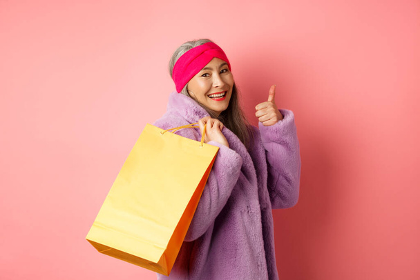 Elegante asiático sênior mulher vai às compras, transportar saco de papel no ombro e mostrando polegar para cima, recomendando descontos loja, fundo rosa - Foto, Imagem