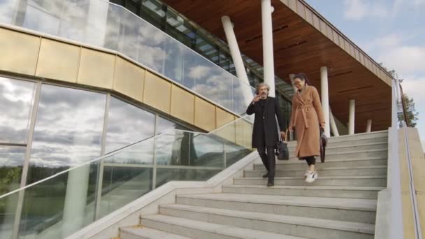 Медленная съемка многонациональной взрослой пары, спускающейся по лестнице, покидающей современное офисное здание во второй половине дня - Кадры, видео