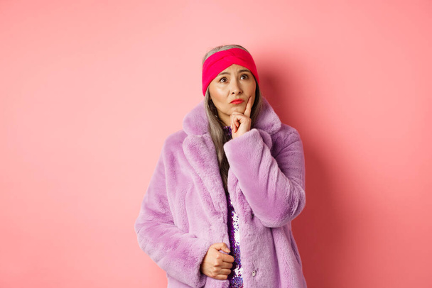 Funky old asian woman looking thoughtful and perplexed, standing in fake fur coat and thinking, making decision, standing on pink background - Zdjęcie, obraz