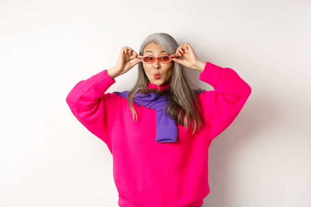 Модная азиатка в солнечных очках, морщинистые губы и удивлённая камерой, удивлённая специальным предложением, стоящая на белом фоне - Фото, изображение