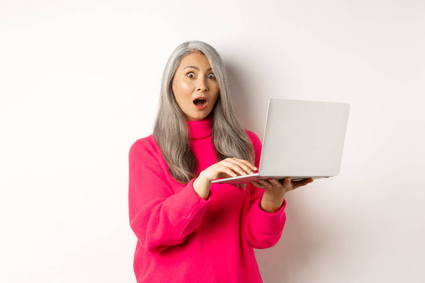 Здивована і вражена азіатська бабуся дивиться шоковані на камеру після читання новин на екрані ноутбука, стоячи над білим тлом
 - Фото, зображення