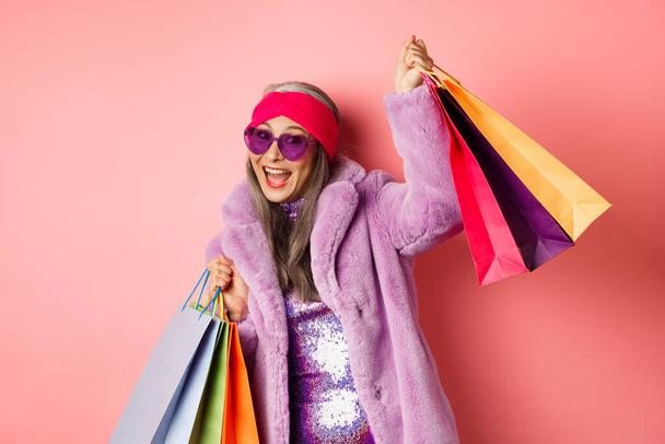 Funky en cool aziatische senior vrouw in modieuze kleding dansen tijdens het winkelen op de verkoop, holding shop papieren tassen en het hebben van plezier, roze achtergrond - Foto, afbeelding