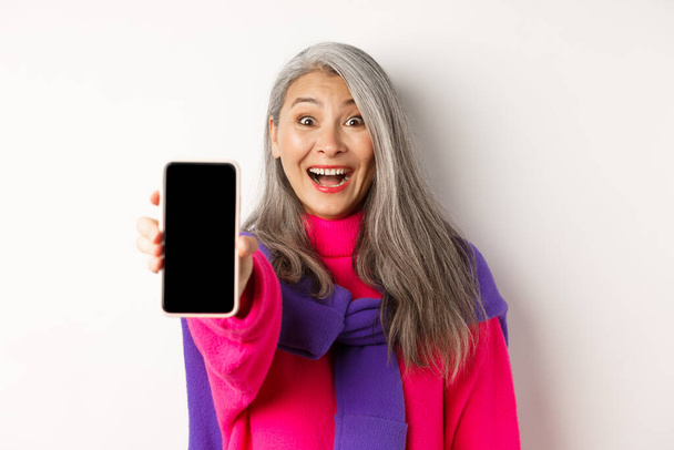 Online nakupování. Detailní záběr stylové asijské starší ženy natahuje ruku s mobilním telefonem, ukazuje prázdné obrazovky smartphone a usmívá se, stojí nad bílým pozadím - Fotografie, Obrázek