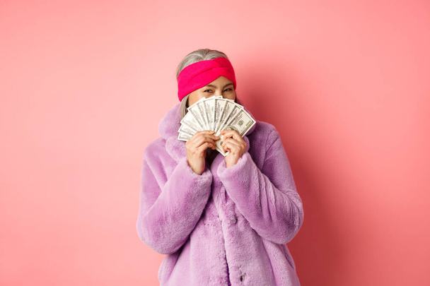 Zakupy i koncepcja mody. Szczęśliwa Azjatka w modnych zimowych ubraniach, trzymając pieniądze i pokazując je przed kamerą, stojąc z dolarami na różowym tle - Zdjęcie, obraz