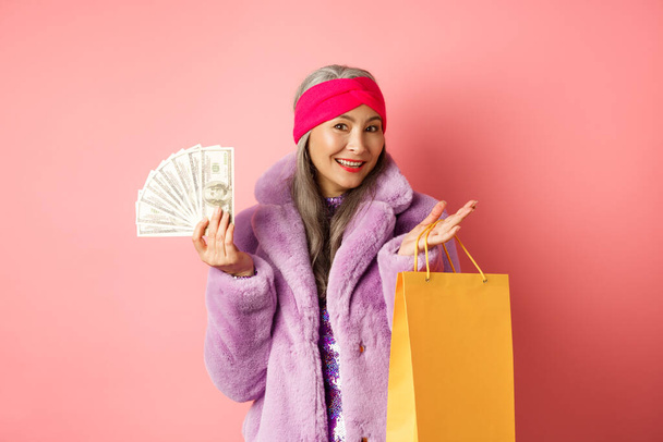 Багата і стильна бабуся збирається на покупки з доларами, тримаючи паперовий пакет і посміхаючись безтурботно, стоїть над рожевим спинкою
 - Фото, зображення
