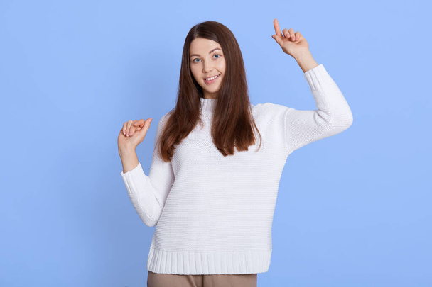 Portrait of optimistic dark haired young woman poses with hands up, dances carefree, wears white jumper, looks at camera, isolated over blue wall, winsome lady celebrates happily. - Фото, зображення