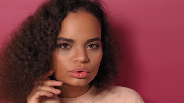 美しいアフリカ系アメリカ人の少女は、ピンクの背景に隔離された桃のシャツを着てカメラをそっと顔に触れている。美しさの概念。4K映像 - 映像、動画