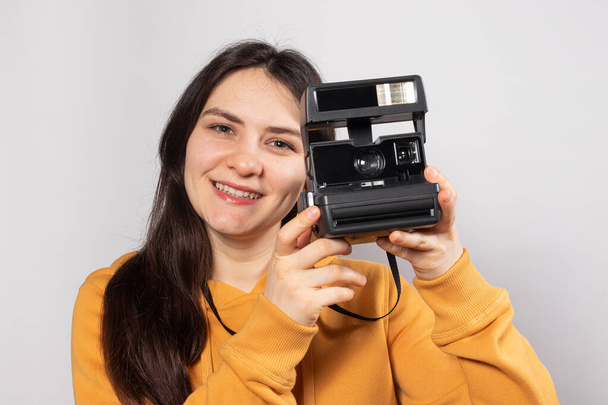 Mooie dame brunette houdt een camera vast en maakt foto 's, glimlachend op een witte achtergrond. Vrouwelijke fotograaf, cursussen en fotografieopleiding. - Foto, afbeelding