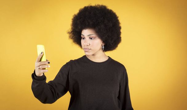 pretty mixed afro woman wearing her smartphone with yellow background - Foto, afbeelding