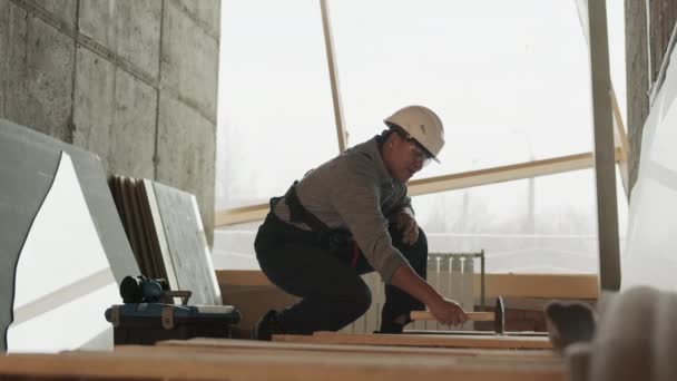 Long shot of Mixed-Race construction worker wearing protective goggles and helmet, squatting in premises under renovation, hammering down on wooden beam - Materiał filmowy, wideo