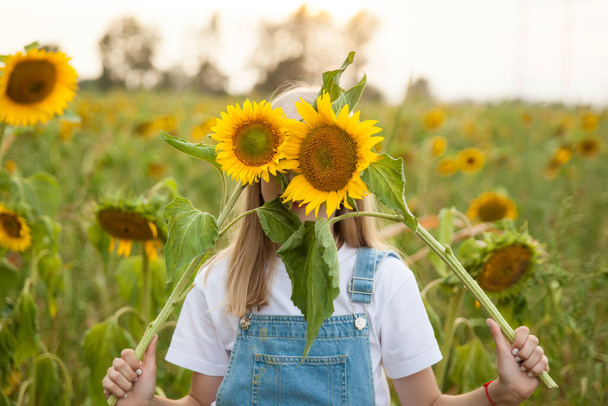 Schöne junge Mädchen in einem blauen Jeansanzug Spaß in einem Feld von Sonnenblumen - Foto, Bild