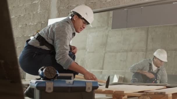 Vollständige Seitenansicht eines Bauarbeiters mit Schutzbrille und Helm, der in renovierten Räumlichkeiten hockt und auf Holzbalken hämmert - Filmmaterial, Video