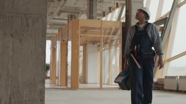 Low angle shot of Mixed-Race female builder wearing protective goggles and helmet, carrying toolbox, looking around in house under construction - Filmati, video