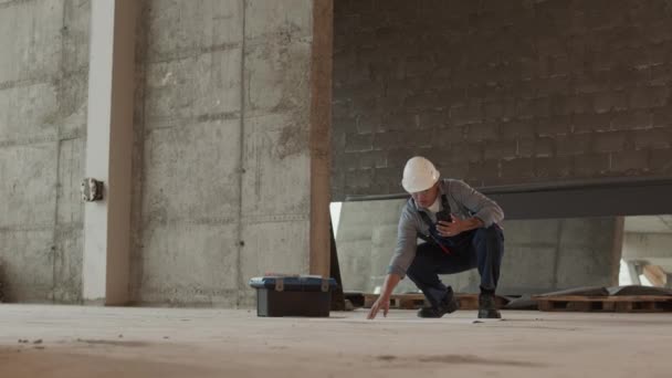 Long shot of professional construction specialist wearing protective goggles and helmet in premises under renovation, holding smartphone in hand, looking at architectural papers on floor - Filmagem, Vídeo