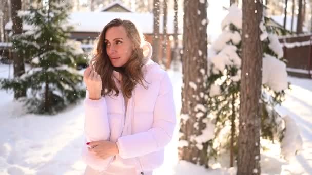 Zimní slunný studený portrét v sněžení krásné mladé ženy v bílém teplé oblečení dolů bunda na dvorku země dřevěný dům na pozadí zeleného smrku borovice pokryté sněhem - Záběry, video