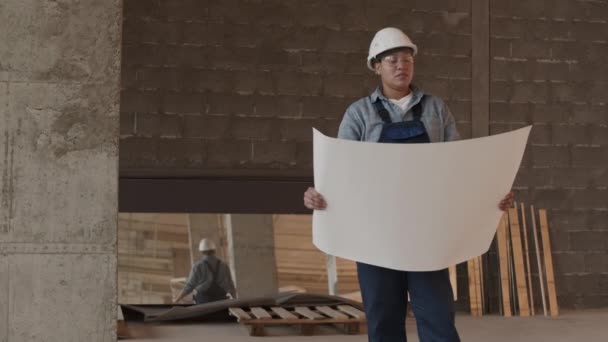 Medium long of Mixed-Race construction specialist wearing protective goggles and helmet, standing in premises under renovation, holding large paper and looking around building - Video