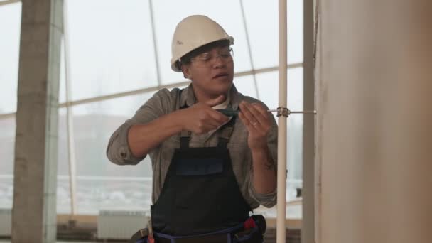 Medium shot of Mixed-Race woman-construction worker wearing helmet and protective goggles, screwing on white plastic pipe with metallic staple to wall - Video