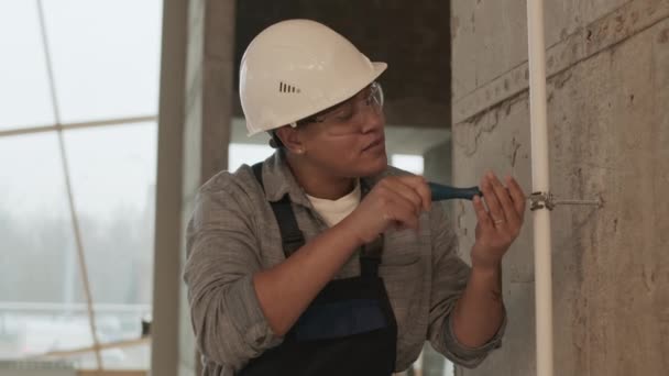 Waist-up of Mixed-Race woman-construction worker wearing helmet and protective goggles, screwing on white plastic pipe with staple to wall - Video