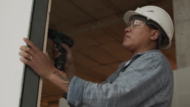 poitrine haute à angle bas d'un ouvrier de la construction mixte portant un casque et des lunettes debout à l'intérieur, à l'aide d'une perceuse murale - Séquence, vidéo