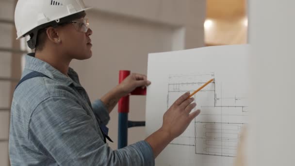 Seitenansicht mittlere Nahaufnahme einer professionellen Architektin mit weißem Helm und Schutzbrille, die in einem Bauobjekt steht und mit folgendem Bleistift auf dem Bauplan überprüft - Filmmaterial, Video