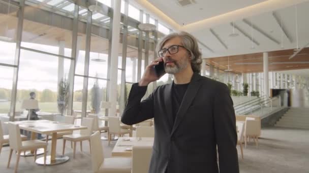 Tracking slow-motion front-view waist-up shot of serious business-like man talking on phone while walking along luxurious restaurant on first floor of modern office building - Footage, Video