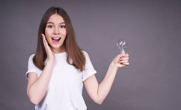 A charming smiling cute young girl in a white casual T-shirt, holds a light bulb in her left hand and looks straight ahead in surprise. Concept, I have an idea. - Foto, immagini
