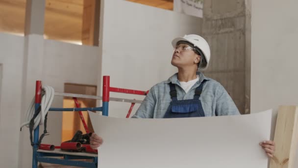 Steadicam shot of Mixed-Race female professional architect wearing white helmet and protective goggles, holding large piece of paper in hands, standing in premises of construction object - Filmati, video