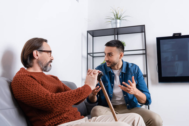 excited hispanic man gesturing while talking to dad in living room - Photo, Image