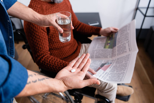 cropped view of man giving water and pill to handicapped father sitting in wheelchair with newspaper, selective focus - Foto, Imagen
