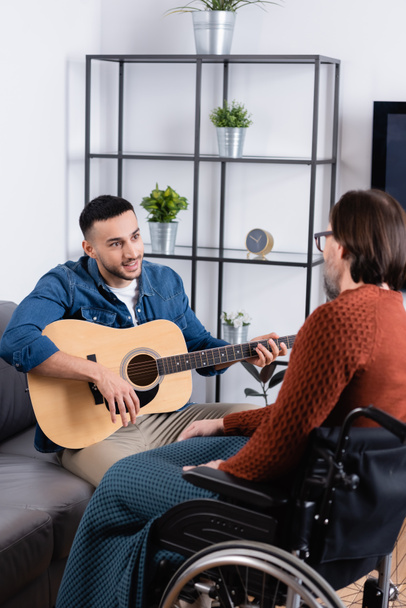 молодий іспаномовний чоловік грає на гітарі біля батька-інваліда на розмитому фоні
 - Фото, зображення